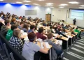 Students attend a lecture in a university auditorium as an instructor delivers a presentation at the front of the classroom.
