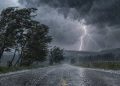 Heavy rain and lightning illuminate a dark, stormy sky as strong winds batter trees along a rural road.