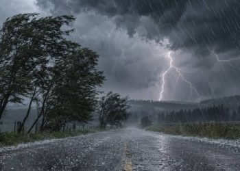 Heavy rain and lightning illuminate a dark, stormy sky as strong winds batter trees along a rural road.