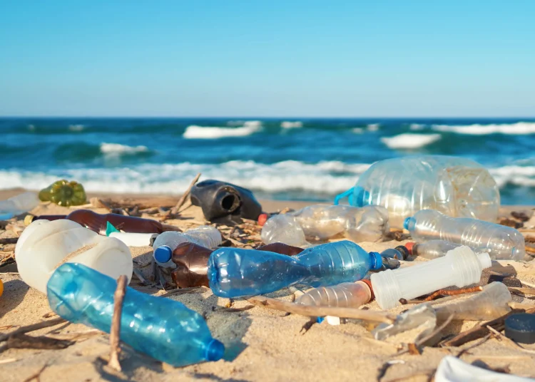 Plastic bottles and other waste litter a sandy beach along the ocean.