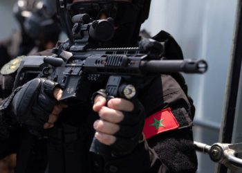 A member of Morocco’s security forces in tactical gear aims a rifle, with the Moroccan flag displayed on his uniform.